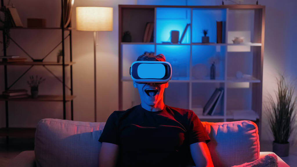 Virtuelle Realität. Männliche Spieler. Cyber-Fortschritt. Erstaunt glücklicher Mann in vr Headset Gefühl Schock der 3D-Welt sitzen dunkel Neon-Licht Home Interieur. - Foto, Bild