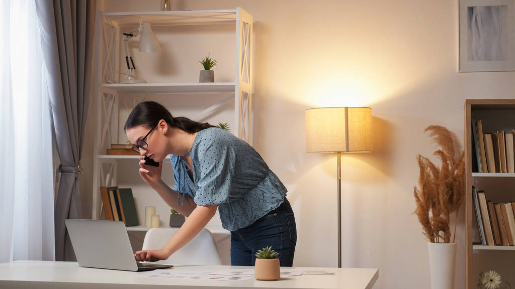 Zarządzanie online. Komunikacja zdalna. Inteligentna, zajęta pracownica rozmawiająca przez telefon o projekcie pracy przy użyciu laptopa we wnętrzu nowoczesnego domu z wolną przestrzenią. - Zdjęcie, obraz