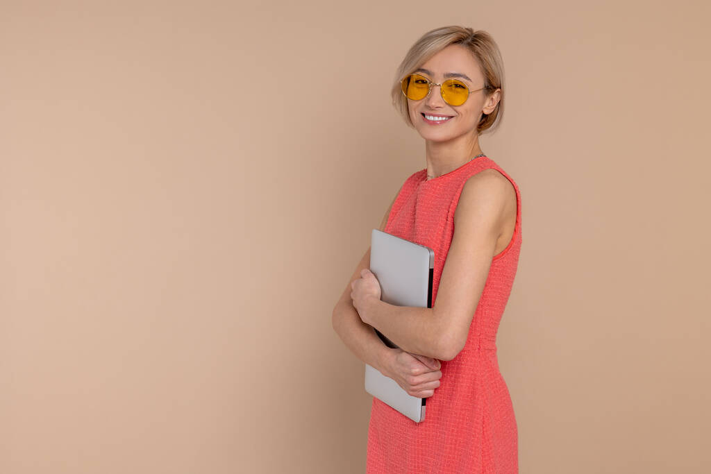 Confiant heureux belle femme en robe et lunettes de soleil tenant ordinateur portable vide espace isolé sur fond beige. - Photo, image