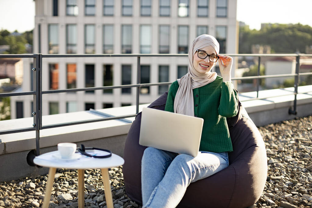 Portret van een moslim dame met een hoofddoek die glimlacht naar de camera terwijl ze 's morgens laptop gebruikt op een panoramisch terras. Professionele ondernemer beheer bedrijf op afstand met behulp van moderne technologieën buiten. - Foto, afbeelding