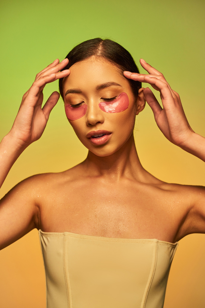 Schönheitsmodell, glühende Haut, junge asiatische Frau mit brünetten Haaren und sauberer Haut posiert und schaut auf grünen Hintergrund, nackte Schultern, feuchtigkeitsspendende Augenklappen, Pflegeprodukt - Foto, Bild