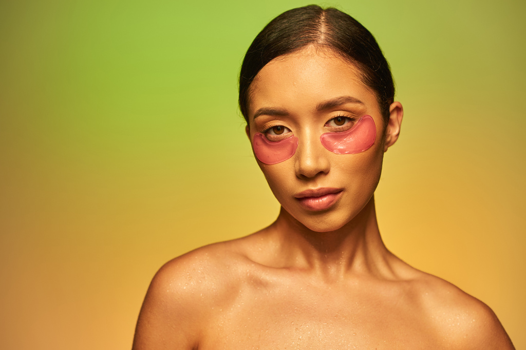 bőrápolási kampány, fiatal ázsiai nő barna hajjal és csupasz vállak pózol és nézi kamera zöld háttér, arcápolás, hidratáló szemtapasz, ragyogó bőr  - Fotó, kép