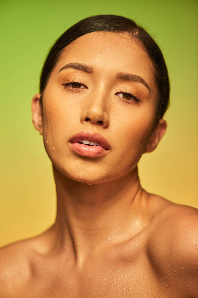 gotas de agua en la cara, primer plano de la joven mujer asiática con la piel mojada mirando a la cámara en el fondo verde, hidratación de la piel, campaña de belleza, perfección, bienestar, tendencia de belleza  - Foto, imagen