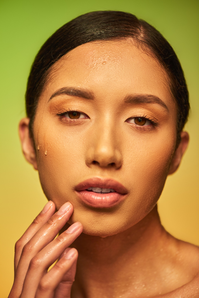 Yüze su damlaları düşer, ıslak ciltli genç Asyalı kadının yeşil arka plandaki kameraya, cilt hidrasyonuna, yüz bakımı kampanyasına, mükemmelliğe, sağlığa, kavramsal konsepte baktığı yakın plan.  - Fotoğraf, Görsel