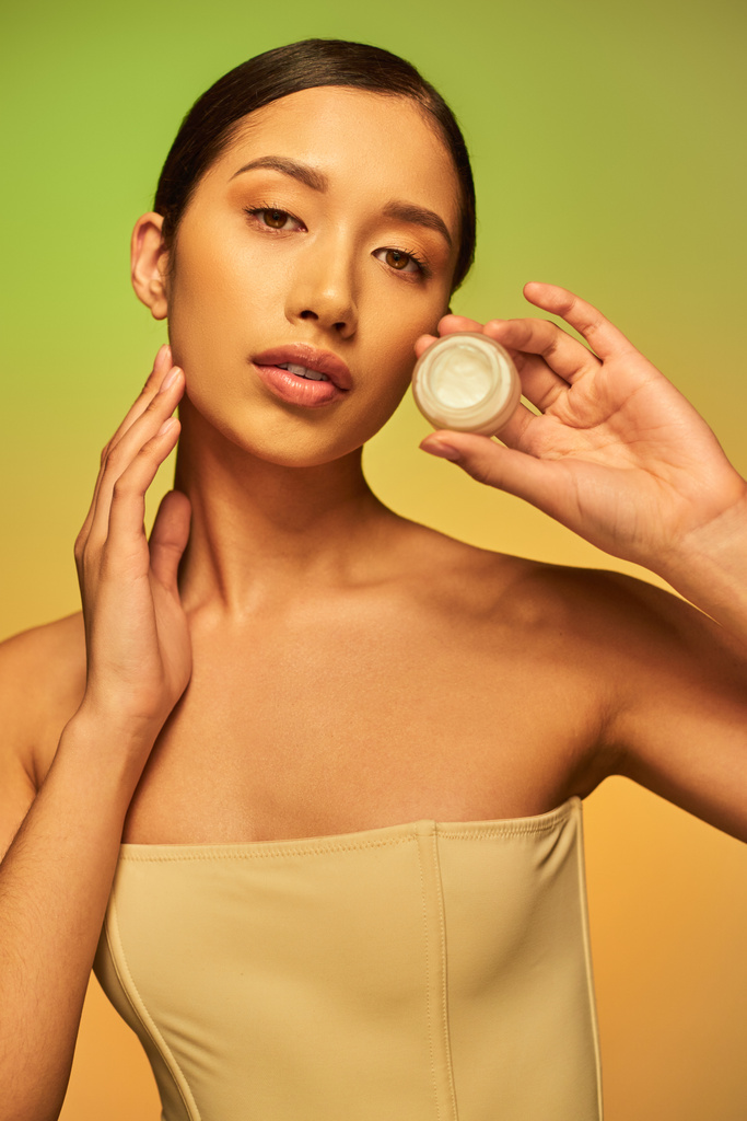 kosmetický výrobek, mladá asijská žena s holými rameny držení kosmetické sklenice s obličejovým krémem na zeleném pozadí, brunetky vlasy, kosmetický průmysl, zářící kůže, péče o pleť koncept  - Fotografie, Obrázek