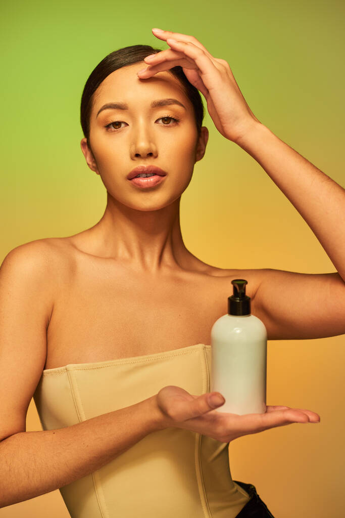 Ürün sunumu, çıplak omuzlu genç Asyalı bir kadın güzellik ürünüyle kozmetik şişesi tutuyor ve yeşil arka plandaki kameraya bakıyor, esmer saçlı, parlak tenli.  - Fotoğraf, Görsel