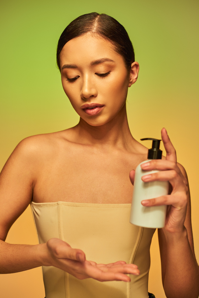 prezentacja produktu, produkt do pielęgnacji skóry, młoda Azjatka z gołymi ramionami trzymająca butelkę kosmetyczną i pozująca na zielonym tle, świecąca skóra, brunetka włosy  - Zdjęcie, obraz