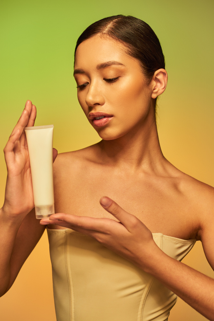 Cilt bakımı, ürün sunumu, genç cilt, çıplak omuzlu genç Asyalı kadın kozmetik tüpü tutuyor ve yeşil arka planda poz veriyor, parlayan cilt, esmer saç  - Fotoğraf, Görsel