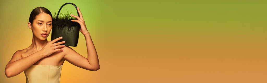 Modewahl, brünette asiatische Frau mit nackten Schultern posiert mit Federhandtasche auf grünem Hintergrund, Steigung, modisches Statement, glühende Haut, natürliche Schönheit, junges Model, Banner - Foto, Bild
