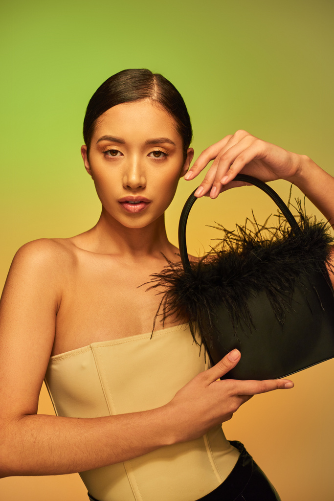 fashion forward, brunette asian woman with bare shoulders posing with feather purse on green background, gradient, fashion statement, glowing skin, natural beauty, young model looking at camera - Photo, Image