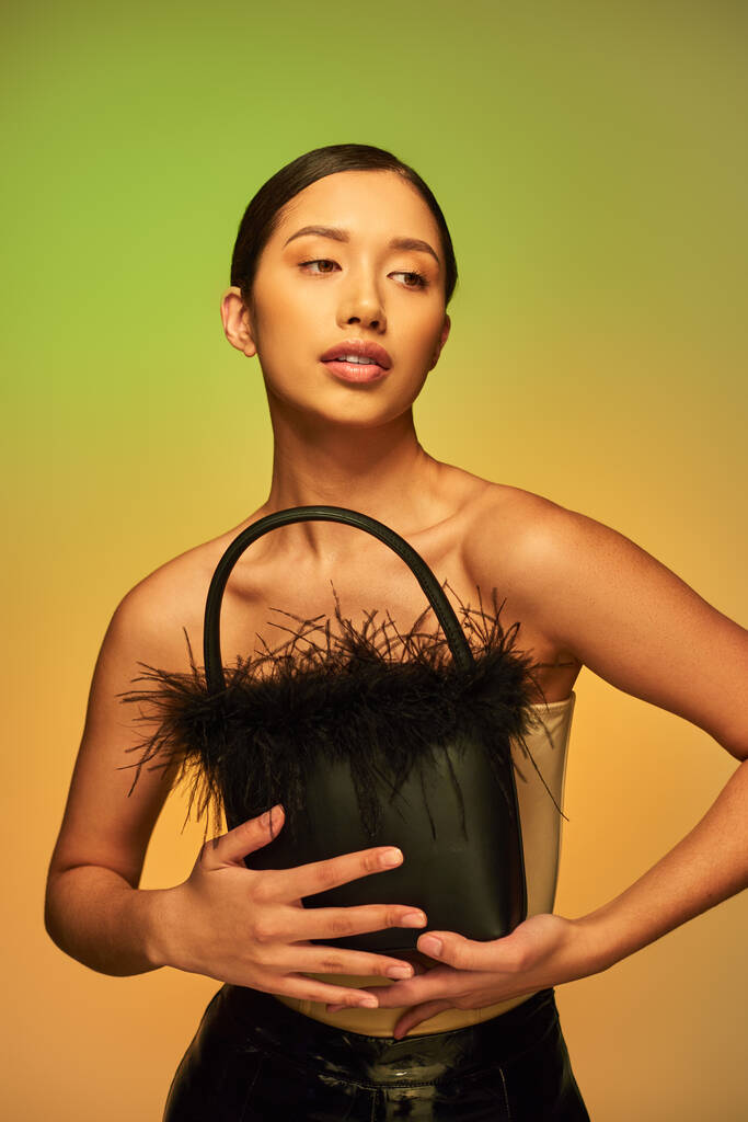 choix de mode, femme asiatique brune aux épaules nues posant avec sac à main en plumes sur fond vert, dégradé, mode vers l'avant, peau éclatante, beauté naturelle, jeune modèle  - Photo, image