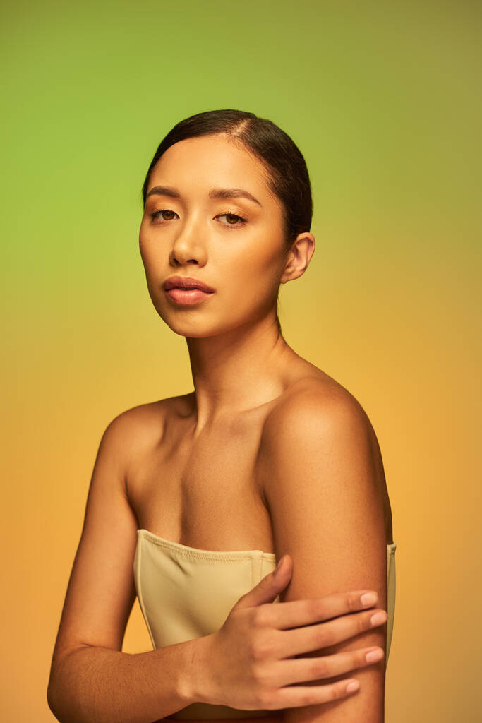 beleza asiática, jovem mulher com cabelo morena e ombros nus posando no fundo gradiente, verde e laranja, cuidados com a pele, pele brilhante, beleza natural, modelo de beleza  - Foto, Imagem