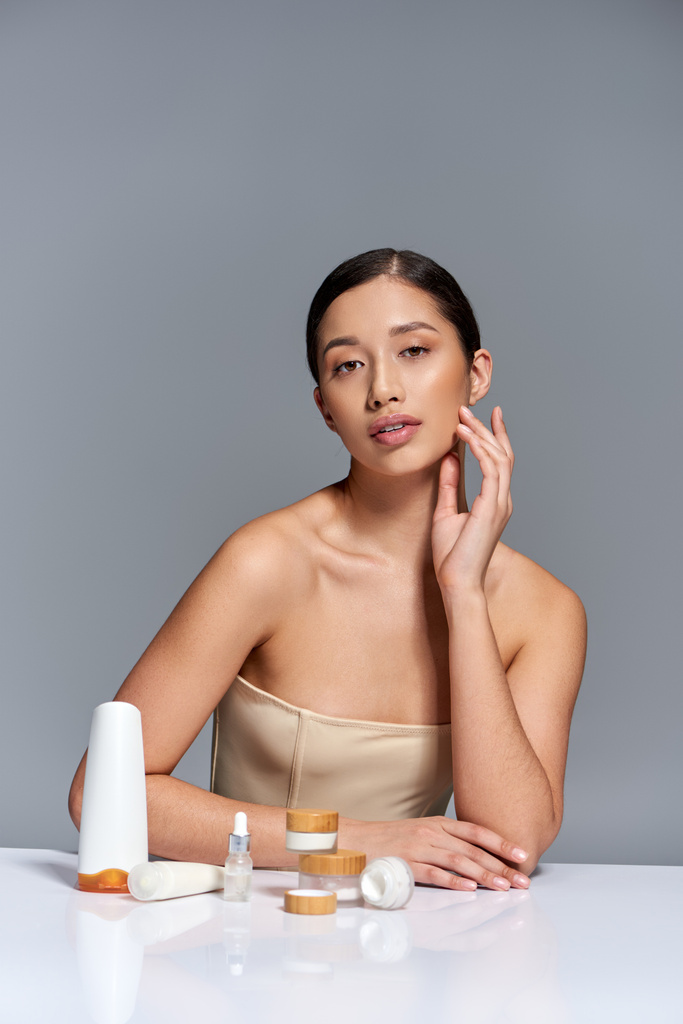 bőrápolás bemutató, fiatal ázsiai modell barna haj pózol közel különböző szépségápolási termékek szürke háttér, izzó és heathy bőr, szépség kampány, arckezelés koncepció  - Fotó, kép