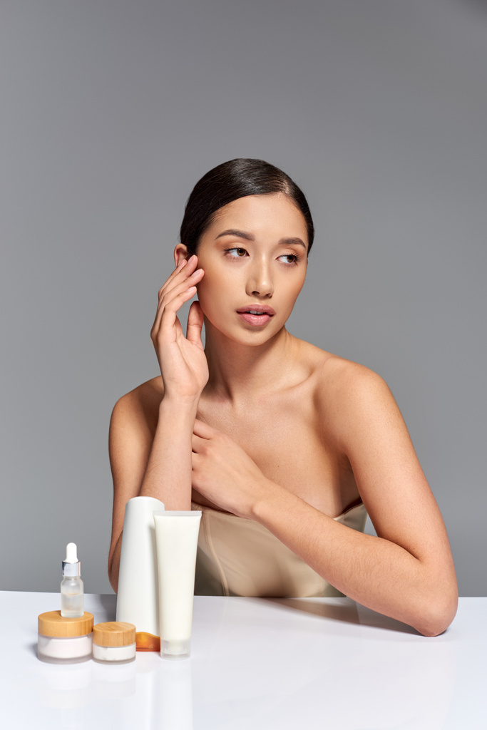 güzellik trendi, cilt bakımı, esmer saçlı genç Asyalı kadın gri arka planda güzellik ürünlerinin yanında poz veriyor, parlayan ve sağlıklı ten, yüz bakımı konsepti, yüz bakımı  - Fotoğraf, Görsel