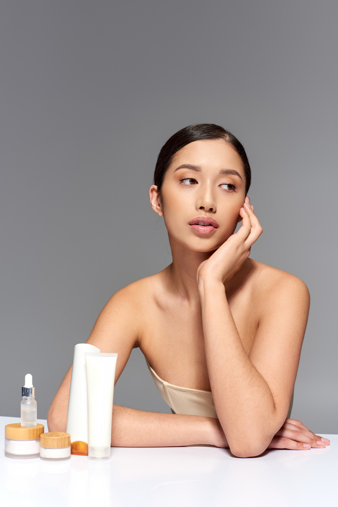 beauty industry, young asian woman with brunette hair posing near beauty products on grey background, glowing and heathy skin, facial treatment concept, facial and skin care  - Photo, Image