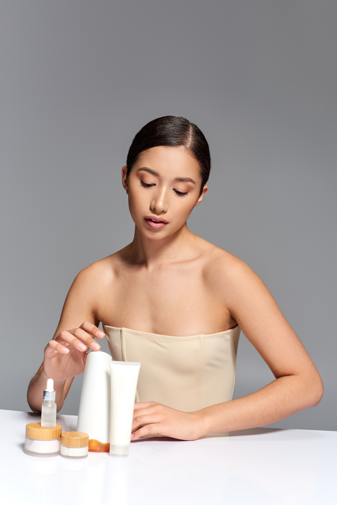 Gesichtsbehandlung, junge asiatische Frau mit brünetten Haaren posiert in der Nähe von Schönheitsprodukten auf grauem Hintergrund, glühende und heide Haut, Gesichtspflegekonzept, asiatische Jugend, Schönheitsprodukte - Foto, Bild