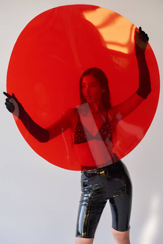 opciones de moda, estilo látex, mujer asiática joven con cabello moreno posando en sujetador y guantes y sosteniendo vidrio rojo en forma redonda sobre fondo gris, opciones de moda, traje elegante, detrás de vidrio - Foto, imagen