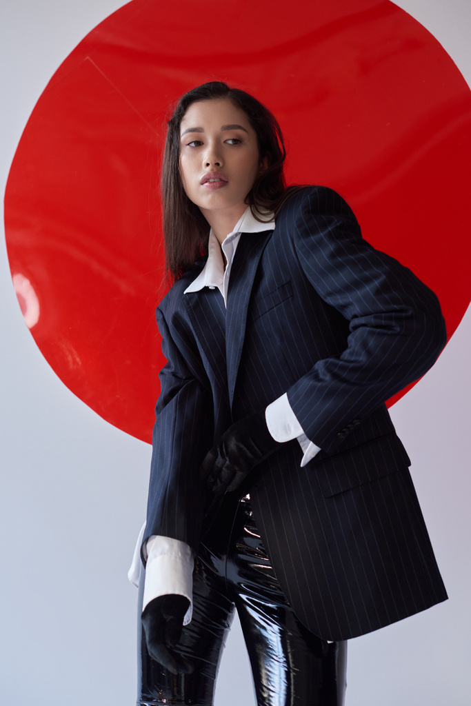 fashion forward, jonge aziatische vrouw in bh, wit shirt en blazer poseren in handschoenen in de buurt van rood rond glas, grijze achtergrond, persoonlijke stijl, latex broek, ondergoed en jas, jeugd  - Foto, afbeelding
