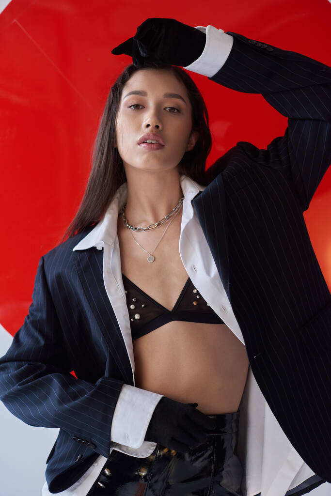 edgy fashion, young asian woman in bra, white shirt and blazer posing in gloves and looking at camera near red round shaped glass, grey background, personal style, underwear and jacket, youth  - Photo, Image