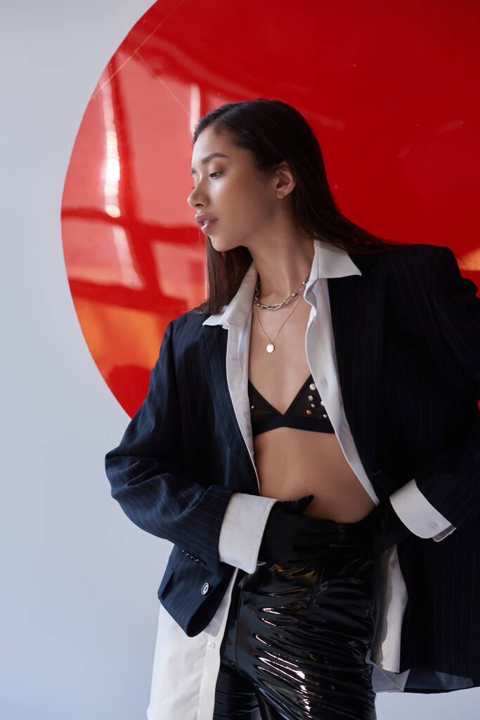 kantige Mode, junge Asiatin in BH, weißem Hemd und Blazer posiert in Latexhose und schwarzen Handschuhen und Latexhosen in der Nähe von rotem Rundglas, grauem Hintergrund, Unterwäsche und Jacke - Foto, Bild