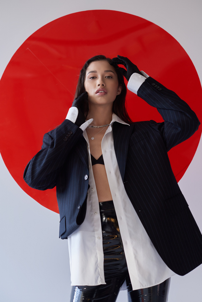 fashion forward, young asian model in bra, white shirt and blazer posing in gloves and latex shorts near red round shaped glass, looking at camera on grey background, personal style, youth  - Photo, Image