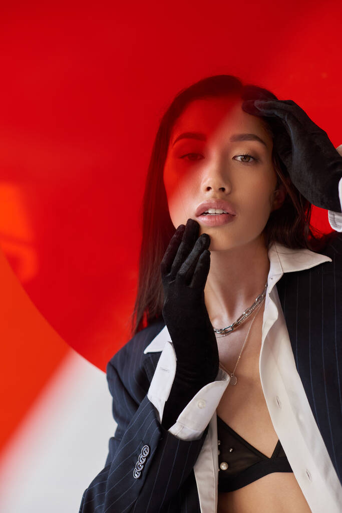 indivíduo moderno, fotografia de moda, jovem modelo asiático em camisa branca e blazer posando em luvas perto de vidro redondo vermelho, fundo cinza, rosto tocante, estilo pessoal, tendência da juventude - Foto, Imagem