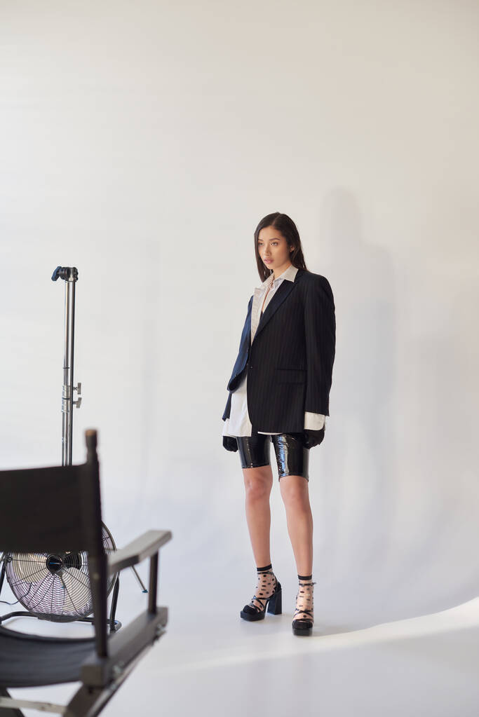 edgy fashion, studio photography, junge asiatische Frau in stylischem Look posiert auf grauem Hintergrund, Blazer, weißes Hemd und Latex-Shorts, neben elektrischem Ventilator stehend, persönlicher Stil, volle Länge  - Foto, Bild