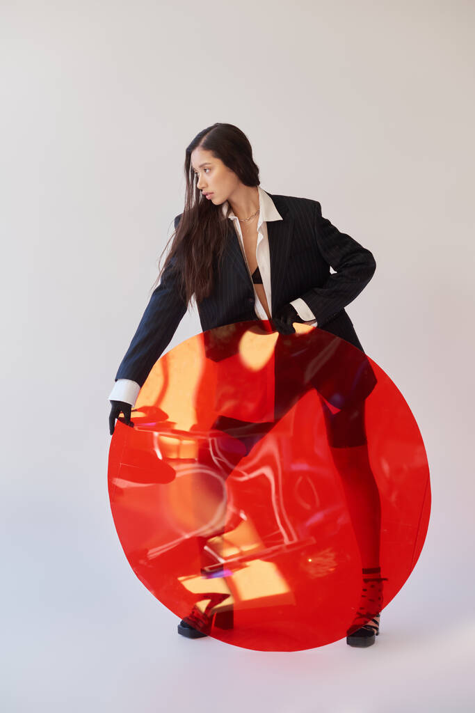 мода и стиль, студийная фотография, молодая женщина в стильной внешности, позирующая возле красного круглого стекла, серый фон, блейзеры и латексные шорты, личный стиль, полная длина  - Фото, изображение