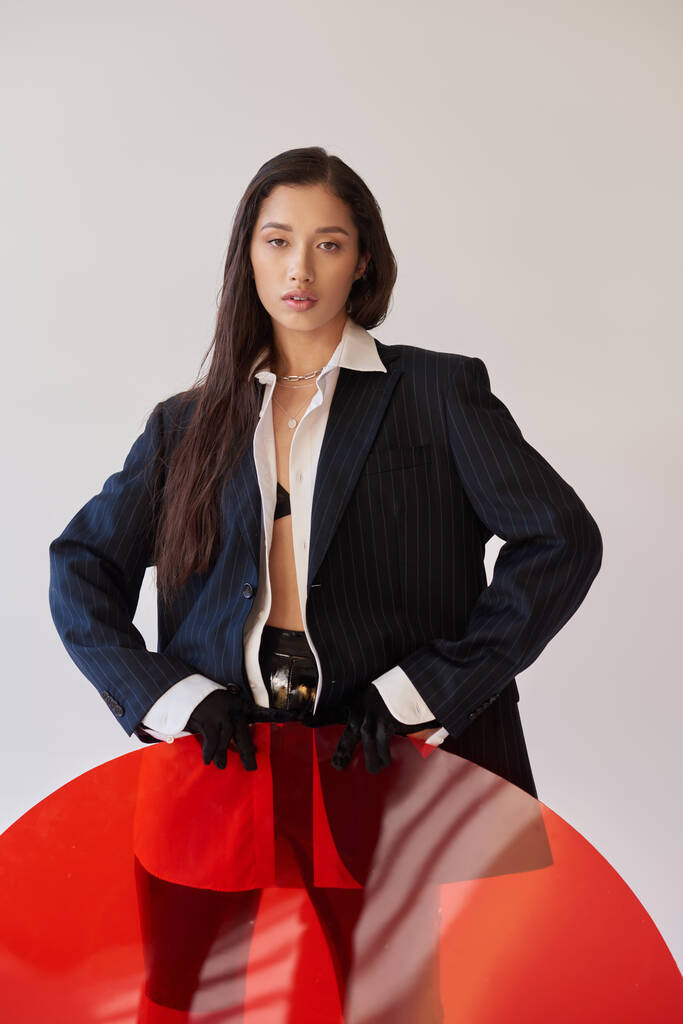 estilo vanguardista, fotografía de estudio, mujer asiática joven en aspecto elegante posando cerca de vidrio redondo rojo, fondo gris, blazer y pantalones cortos de látex, moda juvenil, estilo fresco  - Foto, imagen
