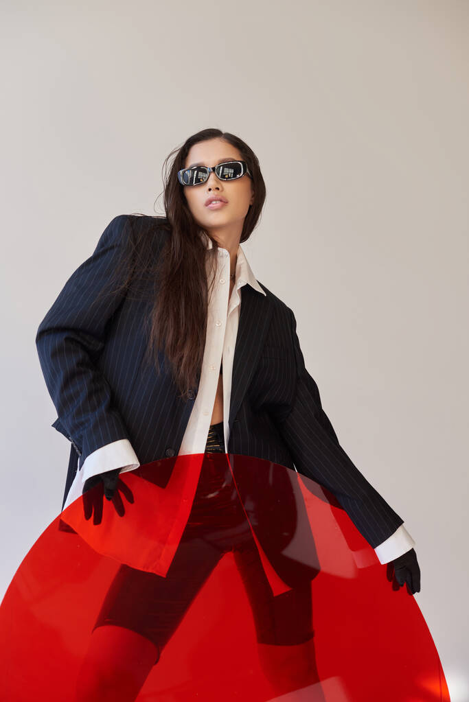 edgy style, studio photography, young asian model in stylish look and sunglasses posing near red round shaped glass, grey background, blazer and latex shorts, youthful fashion, modern woman  - Photo, Image
