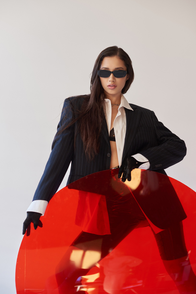 Mode vorne, Studiofotografie, junges asiatisches Model in stylischem Look und Sonnenbrille, die in der Nähe von rotem Rundglas posiert, grauer Hintergrund, Blazer und Latex-Shorts, jugendliche Mode, moderne Frau  - Foto, Bild