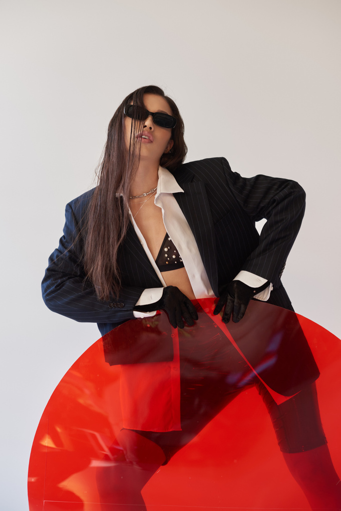 молодая азиатская модель в стильном виде и солнцезащитных очках позирует возле красного круглого стекла, серого фона, пиджака и латексных шорт, молодой моды, современной женщины, острый стиль, студийная фотография  - Фото, изображение