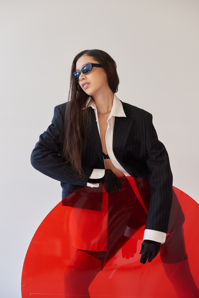 красивая азиатская модель в стильном виде и солнцезащитные очки, позирующие возле красного круглого стекла, серый фон, пиджак и латексные шорты, молодая мода, современная женщина, острый стиль, студийная фотография  - Фото, изображение