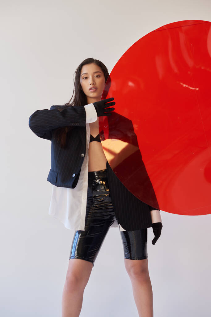 красивая азиатская женщина в модном наряде, держа красное круглое стекло, серый фон, пиджак и черные латексные шорты, молодая модель в перчатках, мода вперед, студийная фотография, концептуальный  - Фото, изображение
