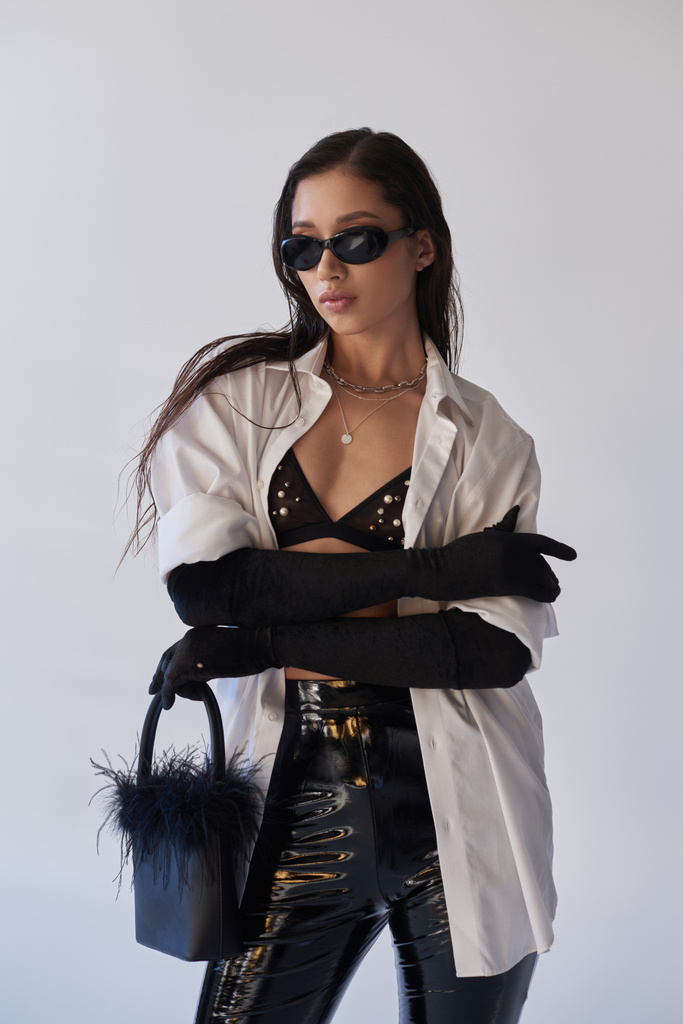 hranatý styl, asijská žena v slunečních brýlích pózuje s opeřenou kabelkou a složené paže na šedém pozadí, mladý model, černé rukavice a bílá košile, latexové šortky, konceptuální, osobní styl  - Fotografie, Obrázek