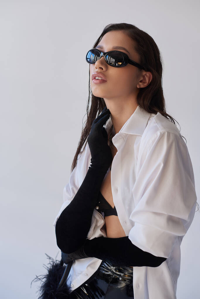 merész stílus, divatos megjelenés, ázsiai nő napszemüvegben pózol tollas kézitáskával szürke háttéren, fiatal modell fekete kesztyűben és fehér ing, divat koncepció, személyes stílus  - Fotó, kép