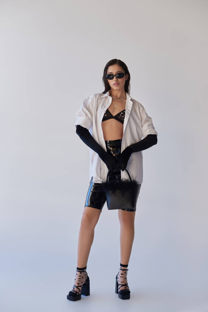 trendy look, fashion statement, brunette asian woman in sunglasses posing with feathered purse on grey background, model in latex shorts, black gloves and white shirt, youth, full length  - Photo, Image