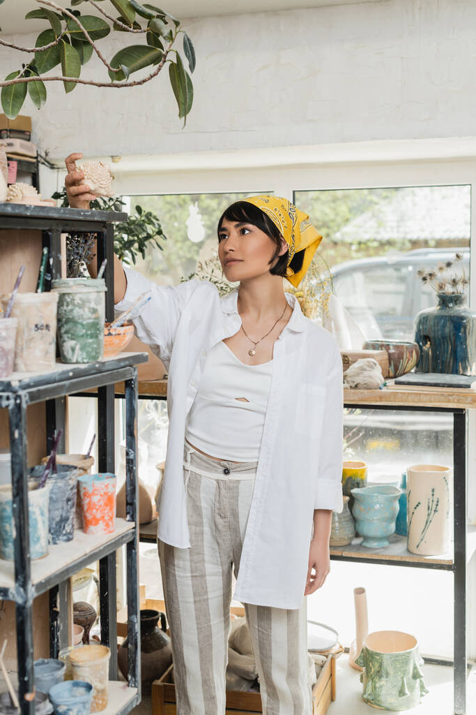 作業着の若いアジア系女性アーティストや棚から粘土製品を取るスカーフ、背景にぼやけた陶芸教室に立っている間、仕事で職人と陶芸スタジオ - 写真・画像