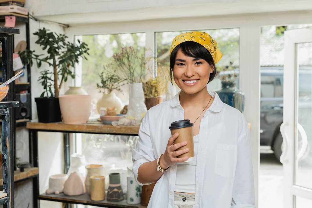 Positive young asian artist in headscarf and workwear looking at camera and holding coffee to go while standing in blurred pottery class at background, pottery workshop with skilled artisan - Photo, Image