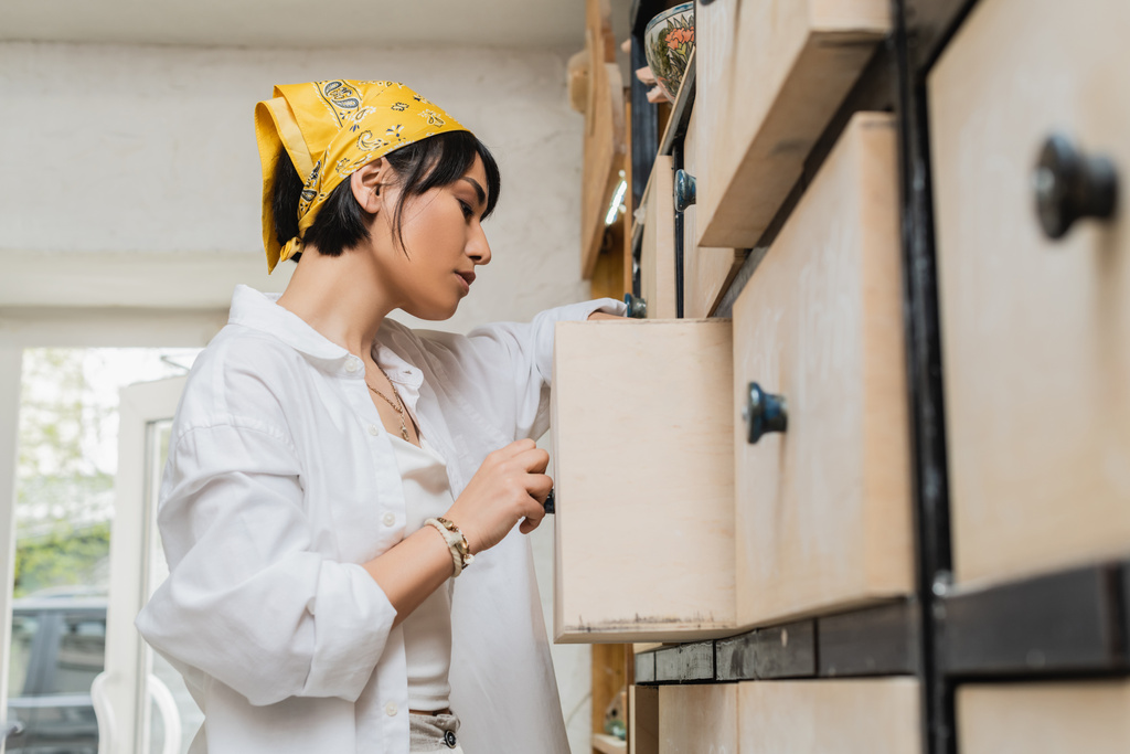 Oldalsó nézet fiatal barna ázsiai női művész munkaruhában és fejkendő nyitó szekrény, miközben dolgozik elmosódott fazekas osztály, kerámia műhely képzett kézműves - Fotó, kép