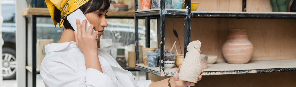 Side view of brunette asian potter in headscarf and workwear talking on smartphone and holding clay sculpture while working in ceramic studio, artisan in pottery studio focusing on creation, banner - Photo, Image