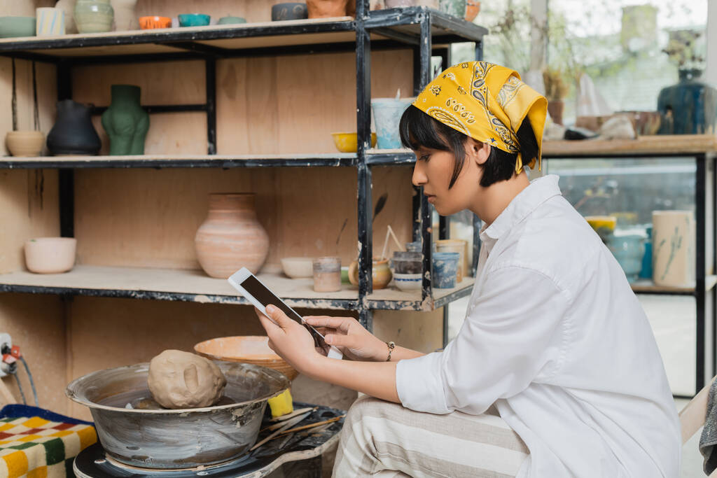 Vue latérale de la jeune artiste féminine asiatique brune en foulard et vêtements de travail à l'aide d'une tablette numérique avec écran blanc alors qu'elle était assise dans un studio en céramique en arrière-plan, artisanat dans la fabrication de poterie - Photo, image