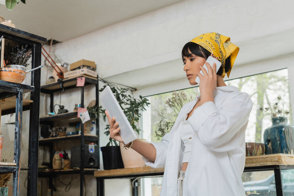Fiatal barna ázsiai női fazekas fejkendőben és munkaruhában beszél okostelefonon és digitális táblagépet tart homályos kerámia műhelyben, kreatív folyamat kerámia készítés - Fotó, kép