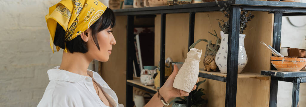 Vista lateral da jovem morena asiática oleiro feminino em véu e workwear segurando escultura de argila perto de prateleiras em oficina de cerâmica no fundo, processo criativo de fabricação de cerâmica, banner  - Foto, Imagem