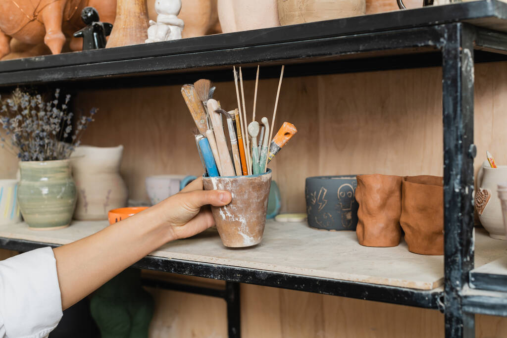 Обрезанный вид молодой ремесленницы с полки с глиняными изделиями и размытыми цветами лаванды на заднем плане в керамической мастерской, творческий процесс изготовления керамики - Фото, изображение
