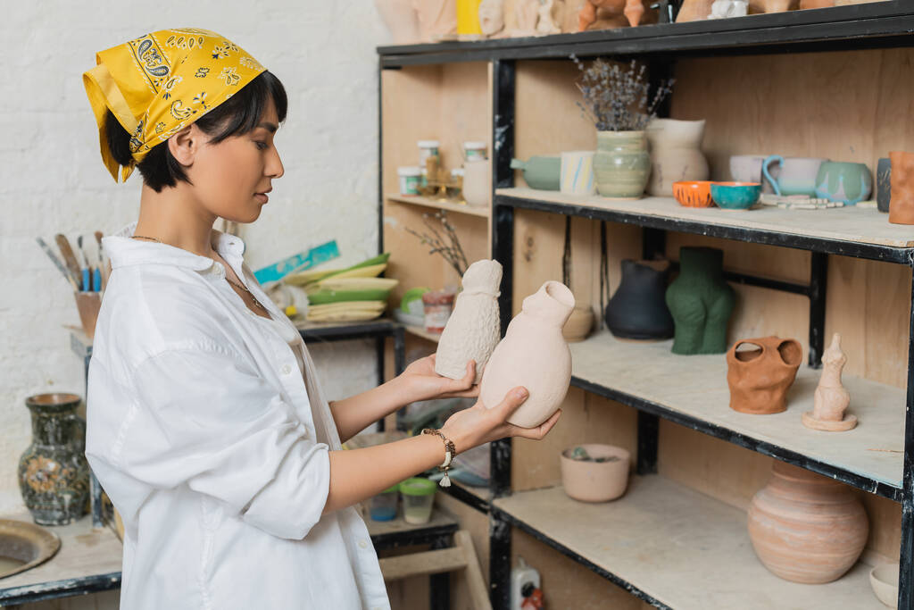 Vista lateral do jovem asiático oleiro feminino em véu e workwear segurando esculturas de argila enquanto estava perto de rack em oficina de cerâmica, cena de estúdio de cerâmica com artesanato qualificado - Foto, Imagem