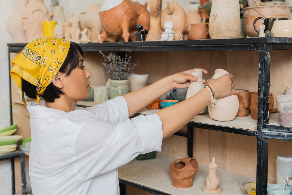 Zijaanzicht van jonge Aziatische vrouwelijke pottenbakker in hoofddoek en werkkleding zetten klei product op rek tijdens het werken in keramische werkplaats, aardewerk studio scène met geschoolde ambachtelijke - Foto, afbeelding