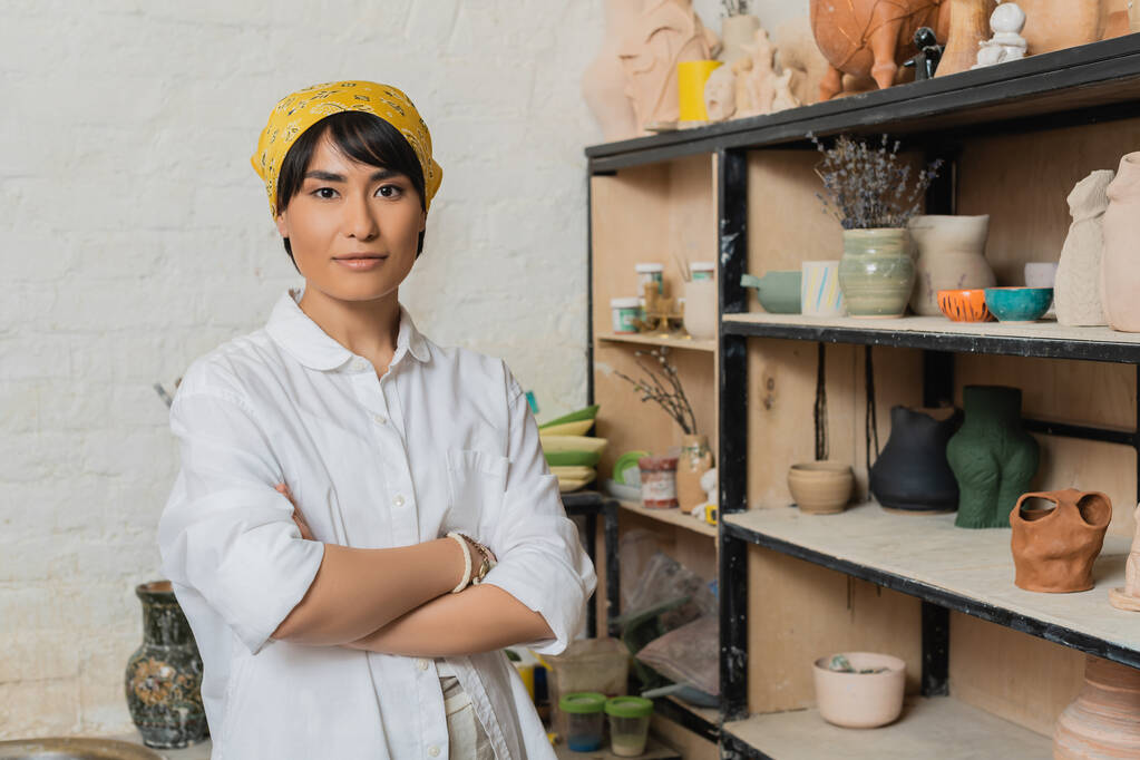 Portré fiatal ázsiai női kézműves fejkendő és munkaruha keresztező karok és nézi kamera közelében állvány agyag szobrok a háttérben, fazekas stúdió jelenet képzett kézműves - Fotó, kép