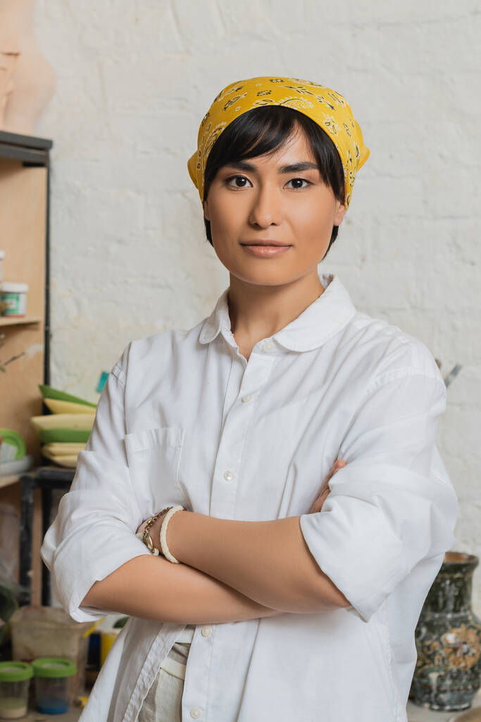 Portrait de jeune potière asiatique en foulard et vêtements de travail croisant les bras et regardant la caméra debout dans un atelier de céramique, scène de studio de poterie avec un artisan qualifié - Photo, image