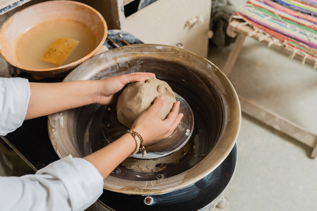 Wysoki kąt widzenia młodych kobiet rzemieślnik w odzieży roboczej formowanie gliny podczas pracy z koła ceramiki w pobliżu miski z wodą w warsztacie, ceramika studio scena z wykwalifikowanym rzemieślnikiem - Zdjęcie, obraz